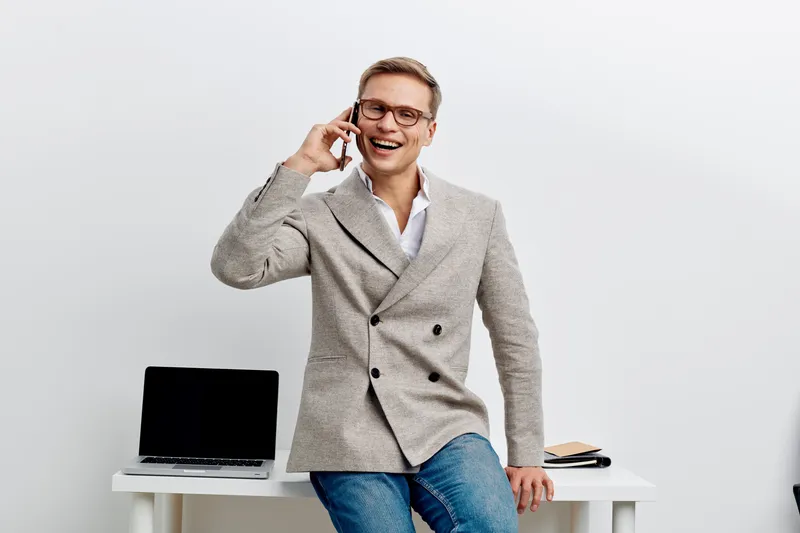 Happy smiling man in business casual attire talking on mobile phone while standing at modern white desk with laptop and notebook in bright office interior. Business communication concept, professional