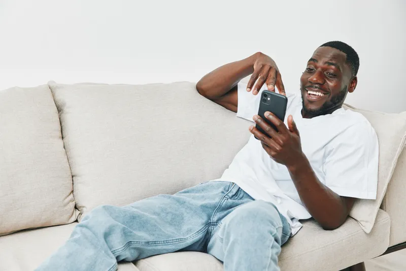 A joyful young Black man smiling while relaxing on a couch and using a smartphone, wearing casual clothing in a modern living room setting