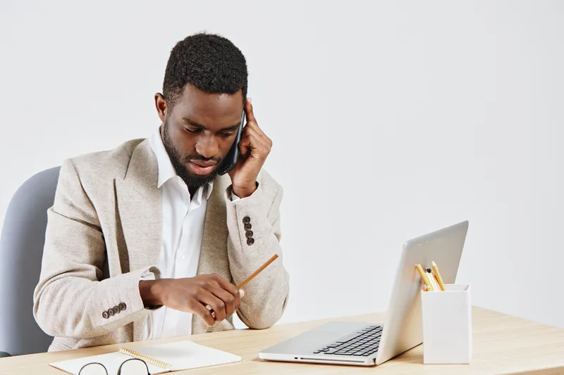 Professional man working with laptop and talking on mobile phone in office setting. Focused, busy, business attire, young adult, African American, modern workspace, preoccupied look, indoor environmen