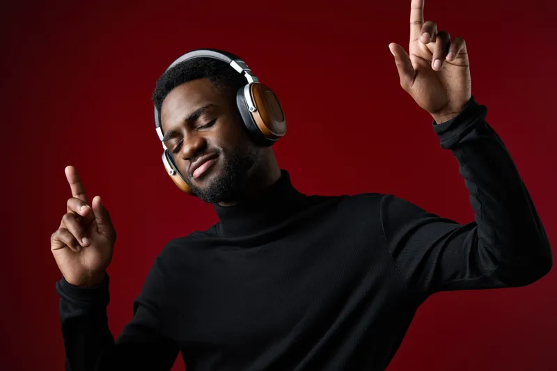 Young Black man enjoying music with headphones, wearing a black turtleneck, dancing against a red background, conveying joy and relaxation