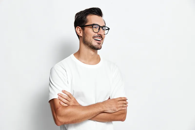 Smiling happy cheerful young man wearing glasses and white t-shirt standing with arms crossed against plain solid light background. People lifestyle concept.
