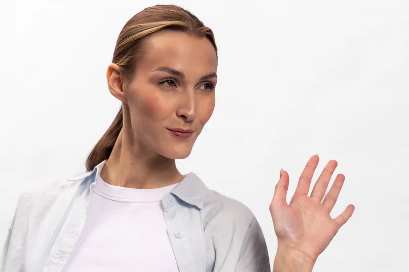 Young woman with light skin and brown hair tied back waves hand with confident expression, wearing white shirt and light jacket. Studio portrait on white background with natural lighting.
