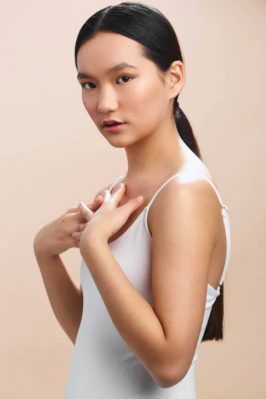 Asian woman in a white sleeveless top stands against a neutral background, presenting a poised and confident expression ideal for lifestyle, fashion, or wellness themes.