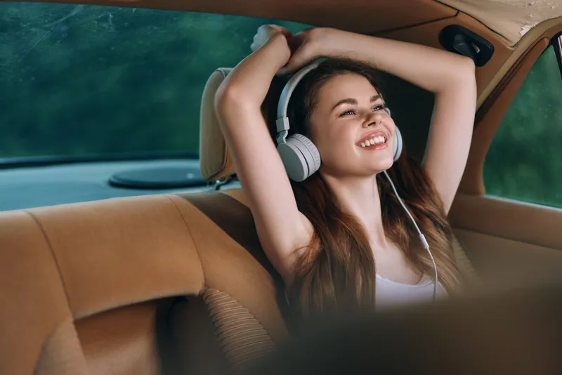 Young woman enjoying music in a car, smiling with headphones, showcasing joy and relaxation in a natural setting.