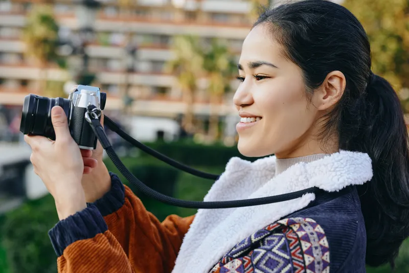 Woman with a camera outdoors, smiling and focused, wearing a warm sweater and scarf, capturing a moment in bright daylight, natural backdrop, candid photography mood.