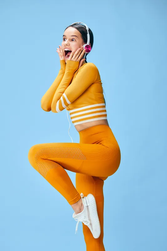 Fitness minded woman in bright orange athletic wear jumps joyfully against a blue backdrop, wearing headphones, capturing a playful, high-energy moment ideal for sport and wellness campaigns.