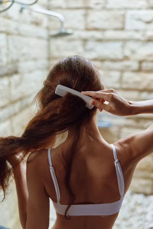 Woman combing long hair indoors near stone wall, soft natural light highlighting brown hair texture and smooth skin in a calm morning routine setting.