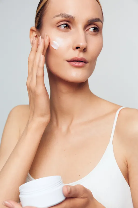 Young woman applies moisturizing cream on clean face with natural skin tone isolated on white background wearing white tank top for skincare beauty routine at home.