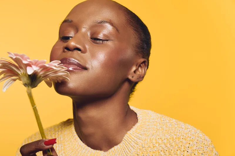 Beautiful dark skinned African woman with closed eyes smelling a flower. Yellow background and sweater create warm mood. Concept of natural beauty, skincare, and wellness.