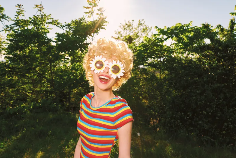 Joyful woman outdoors, wearing a striped rainbow shirt and daisy sunglasses, sunlit moment with relaxed posture amid lush greenery, retro film color mood natural light highlights textures.