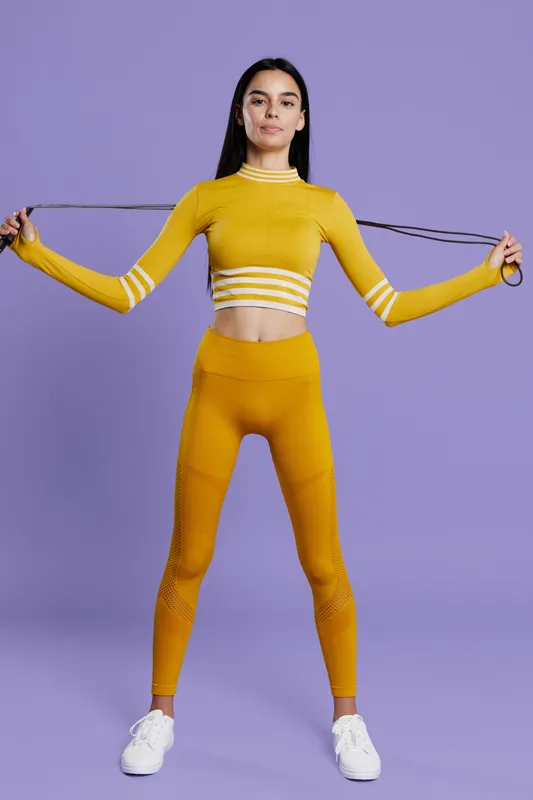 Woman sport fitness dressed in yellow activewear stands against a purple background holding a resistance band, displaying strength, focus, and healthy lifestyle in a modern gym outfit.
