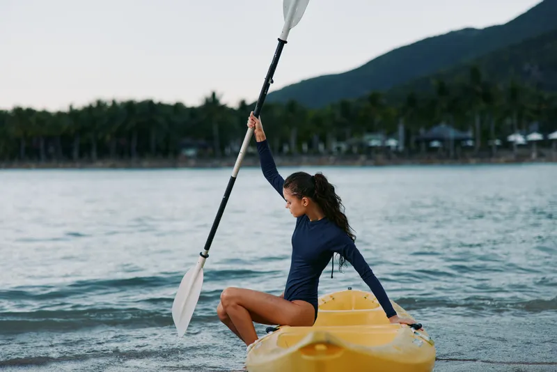 A young woman in a dark blue wetsuit sits on a yellow kayak, raising a paddle as calm water and distant palm trees frame a coastal scene.