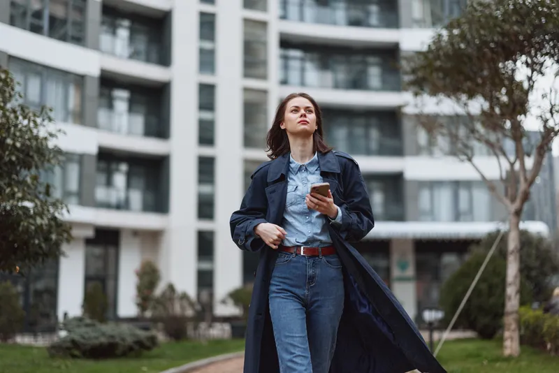 One young woman walks through an urban courtyard wearing a denim jacket and jeans, looking up while holding a smartphone, exuding confidence and purposeful motion today.