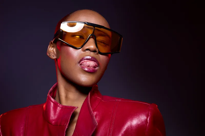 Confident dark skinned African woman wearing oversized yellow sunglasses and red leather jacket poses with tongue out on purple background. Fashion portrait with bold attitude and style.