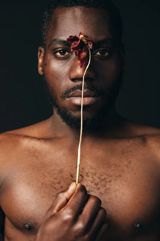 Black man with bare chest holding dried flower in front of face, creative conceptual portrait with visual style, artistic expression and emotional mood on dark background.