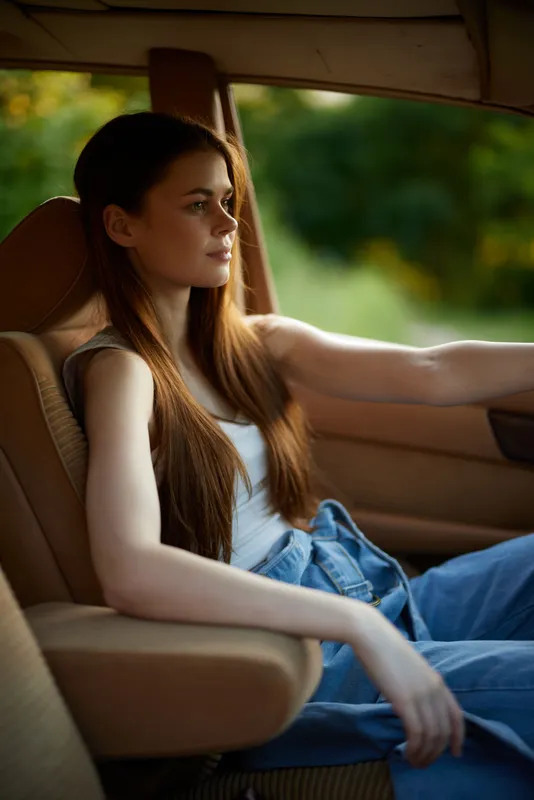 Young woman driving a car, dressed casually in a tank top and jeans, with long hair flowing, showcasing confidence and serenity in a lush green background.