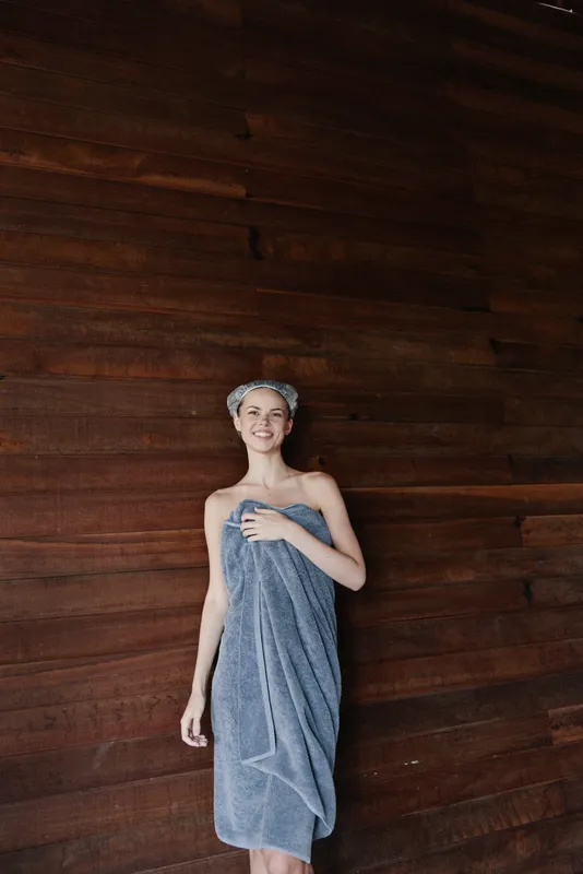 smiling woman wrapped in gray towel standing indoors near wooden wall with relaxed pose, natural light, cozy atmosphere, calm and fresh mood