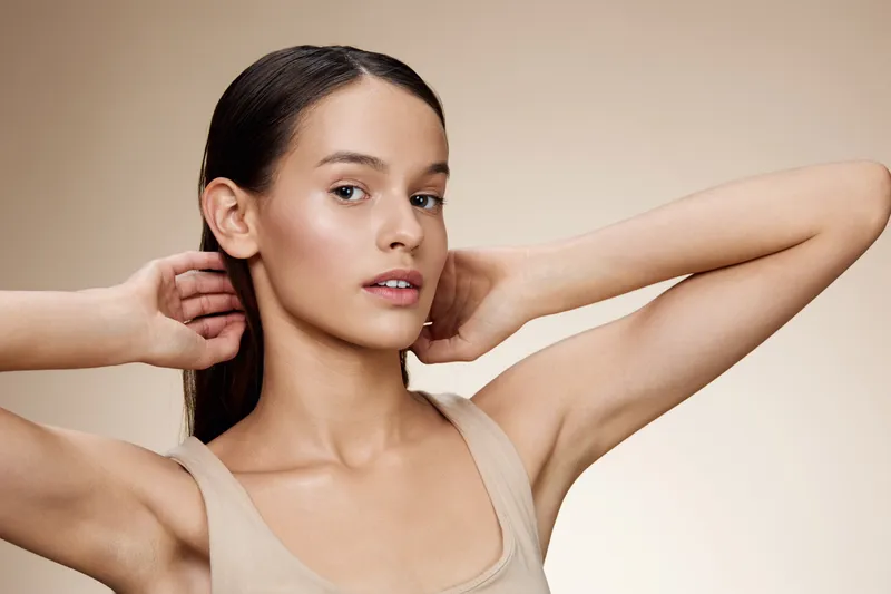 A young woman with long hair wearing a neutral toned sleeveless top, posing confidently with a serene expression against a soft beige background