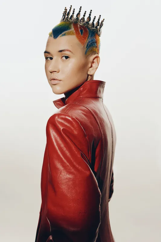 Woman with colorful short hair and artistic crown wearing a red leather jacket, showcasing edgy fashion and bold self-expression in a minimalistic studio setting.