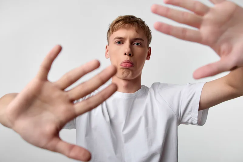Dynamic close-up of a young man wearing a white shirt, reaching hands in front, playful expression with tongue out, studio setting, candid energy, bold composition