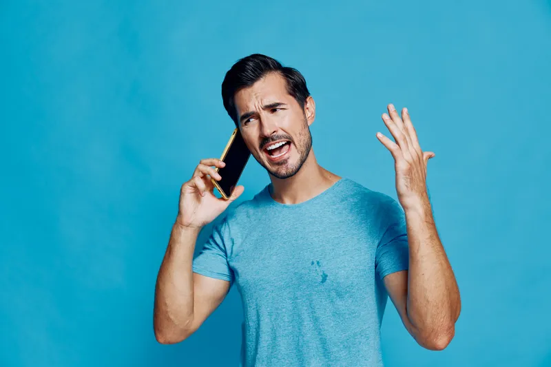 Energetic man talking on smartphone with raised hand, expressing frustration or excitement, wearing a blue t-shirt against a solid blue background.