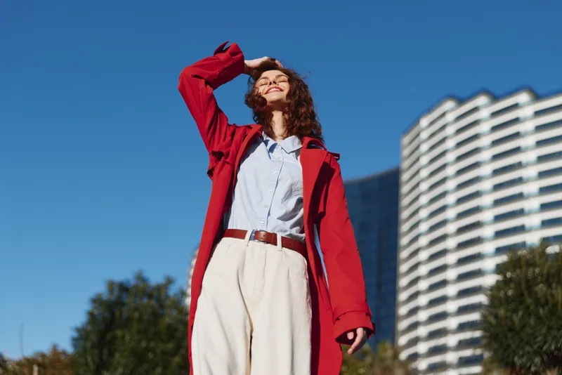 Young woman in a red coat stands outdoors under a blue sky, shielding her eyes with one hand. Fashionable casual look conveys confidence and sunshine.