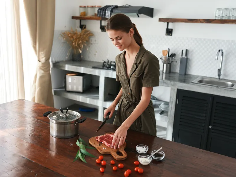 Женщина на кухне улыбается, готовит ужин из мяса и овощей на деревянном столе на фоне стильной кухни, образ жизни