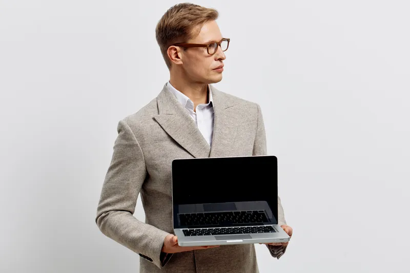 Professional man in smart casual attire holding a laptop computer, standing against a plain white background, studio shot. Business technology concept