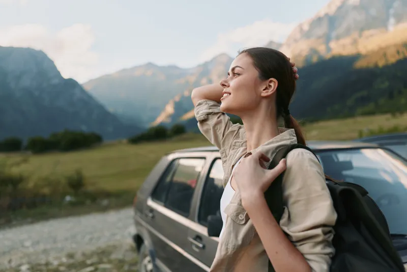 A young woman in casual clothes stands near a car in a mountain landscape, stretching and enjoying fresh air. Concept of lifestyle, travel, and outdoor relaxation.