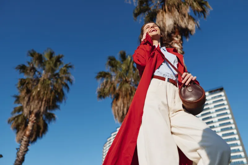 Bold woman in a red coat and light trousers walks outdoors, holding a brown bag under a clear blue sky with palm trees and modern buildings.