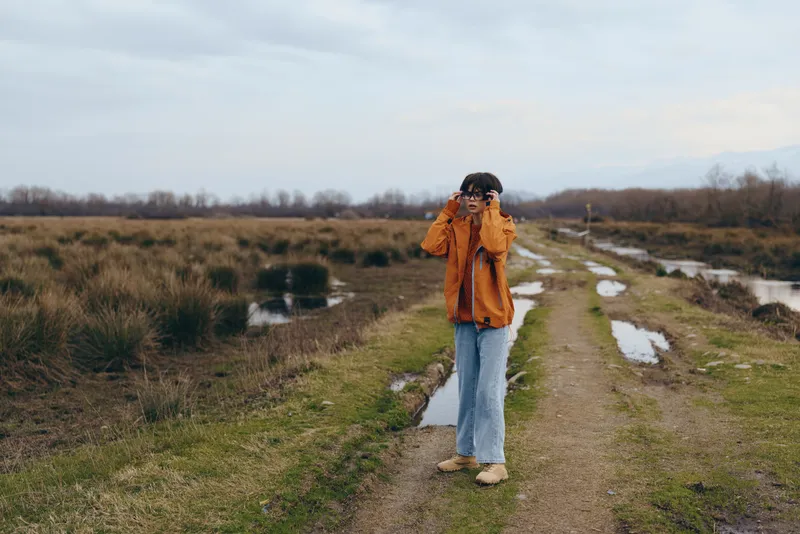 Woman lifestyle hiking trail outdoors nature jacket jeans travel, she walks alone on a dirt path past fields and winter grass, scenic countryside landscape for calm adventure outdoors