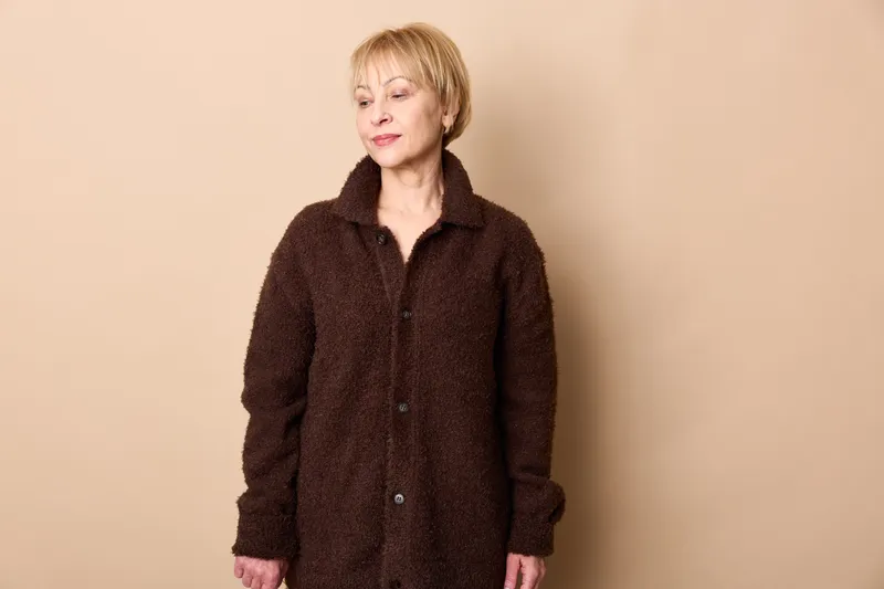 Senior woman in dark coat, standing indoors in a beige studio portrait, calm expression, soft lighting, minimal fashion concept for mature style