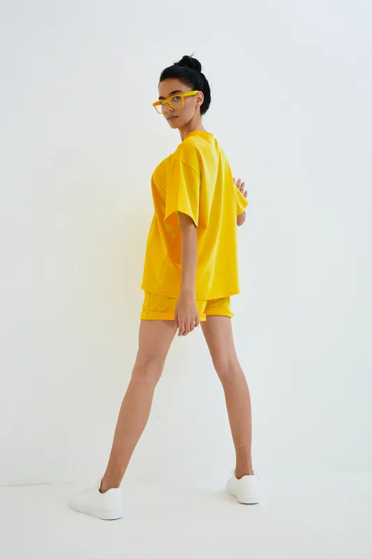 Young woman wearing bright yellow casual outfit and matching glasses, walking confidently against a minimalist white background with white sneakers.