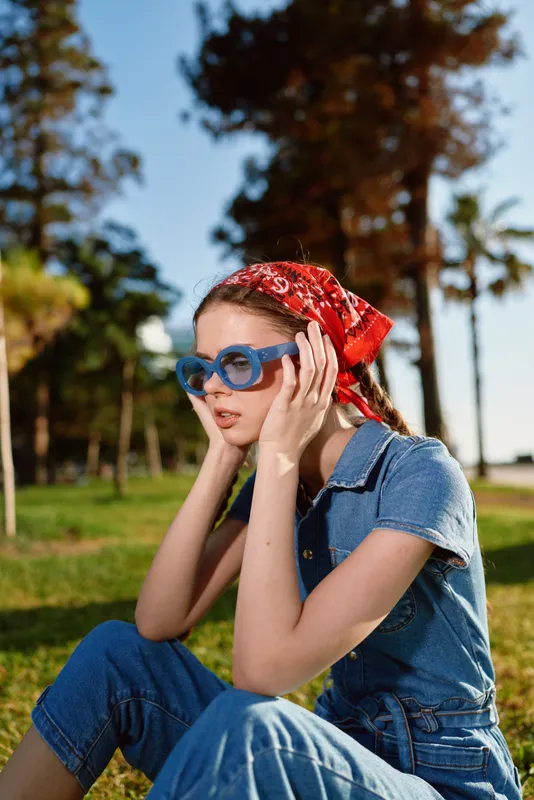 young woman wearing blue denim outfit and red bandana sitting outdoors in sunny park with trees and grass background