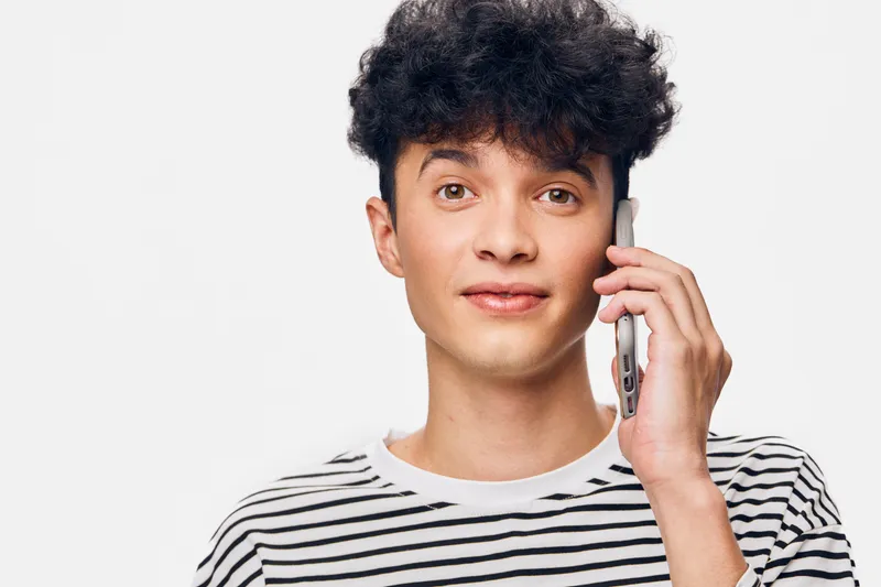 young man using smartphone with curious expression, wearing striped shirt against white background, modern casual style