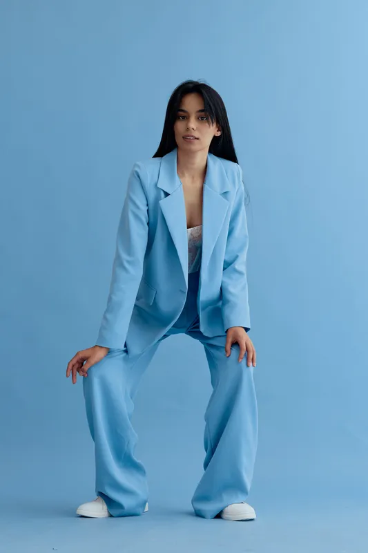 Woman in light blue pantsuit posing confidently against plain blue background, wearing white shoes and relaxed hairstyle, showcasing modern fashion style.