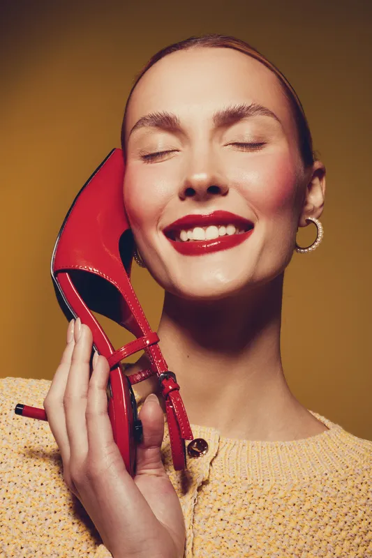Smiling woman with closed eyes holds red high heel shoe near face. Bright makeup and cozy sweater create warm mood. Concept for fashion, beauty, and style accessories.