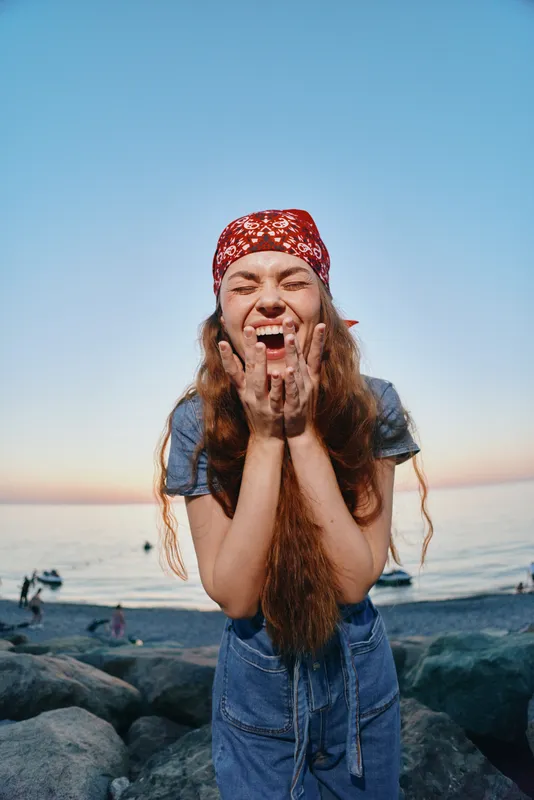 happy woman laughing outdoors near rocky beach with sea background and clear blue sky during sunset, casual outfit and red bandana on head
