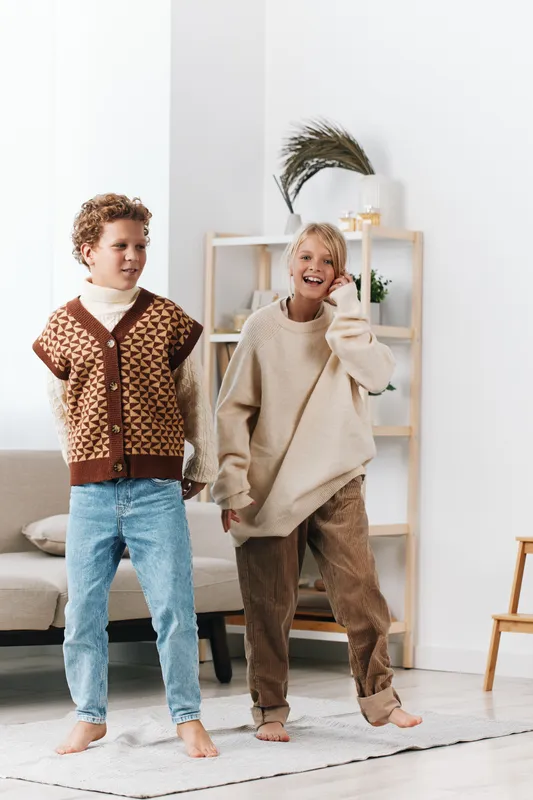 Two children dance indoors in cozy knitwear, smiling as they twirl in a bright living room with soft light, simple decor, and a playful, carefree mood.