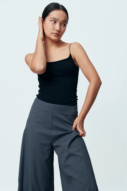 Young woman posing in studio with black tank top and wide gray pants against plain background. Casual fashion, confident model, minimalistic style and modern clothing for portrait photography.