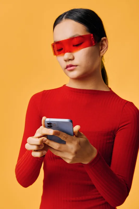 Young woman in red sweater and futuristic red glasses using smartphone for texting with serious expression on orange background for technology and communication concept.