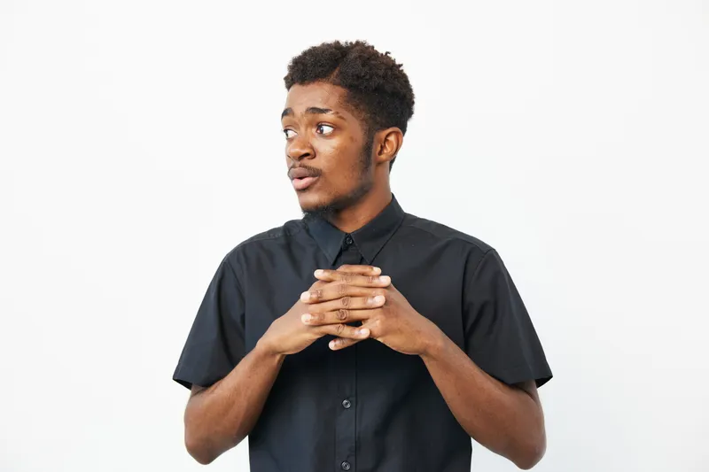 A thoughtful African American man in a black shirt, with an expressive pose that conveys curiosity and engagement. He stands against a clean white backdrop.