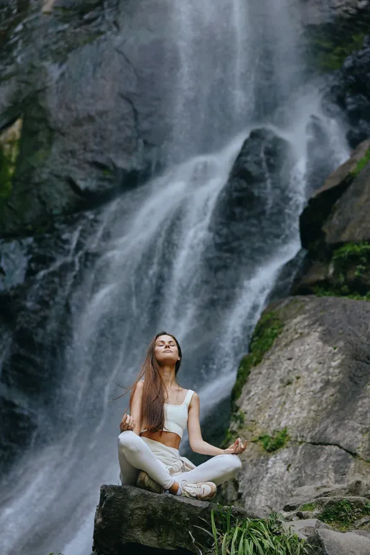 young woman meditating peacefully near a waterfall surrounded by rocks and lush greenery in natural outdoor setting during daytime