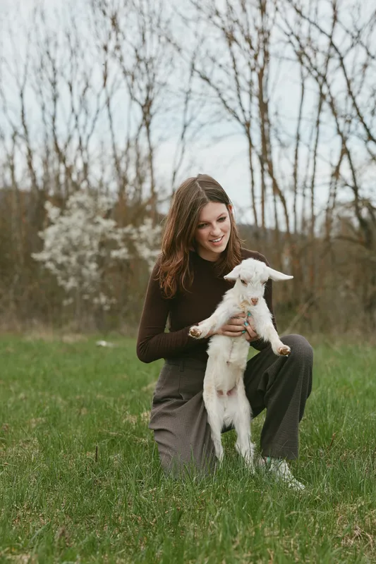 A woman kneels in a green meadow, holding a friendly kid goat, capturing a quiet moment of bond, joy, and rural lifestyle in an open countryside setting.