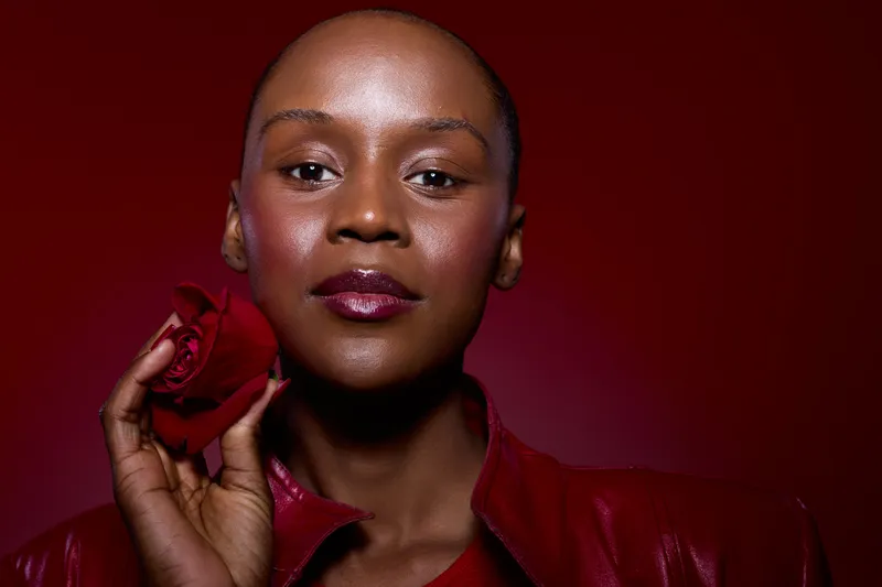 Close-up of a confident dark skinned African woman with short hair holding a red rose near her face against a dark red background. Concept of beauty, elegance, and self-confidence.