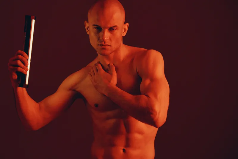 Muscular man holding a light in a dramatic pose, showcasing strength and confidence in a striking red ambiance. Perfect for fitness and artistic themes.