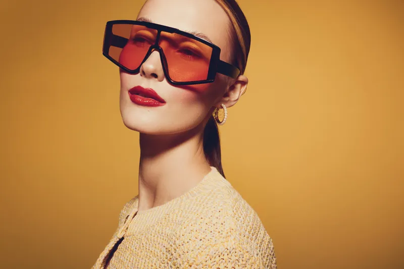 Stylish woman with oversized orange sunglasses and red lipstick poses against yellow background. Fashion portrait with trendy accessories and confident expression.
