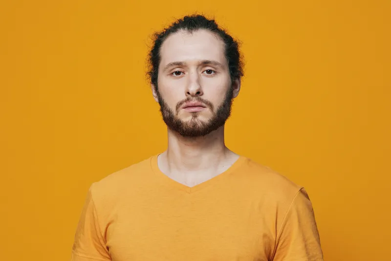 Young confident man with curly hair wearing a yellow shirt, standing against a vibrant orange background, exuding self assuredness and positivity