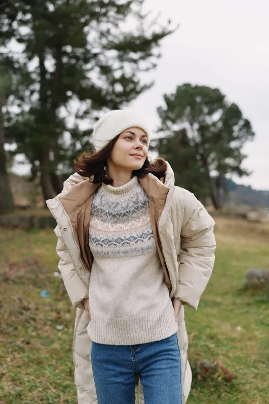 Woman in a cozy winter coat and knit sweater stands outdoors in a grassy field, wearing a hat, with soft trees in the distance