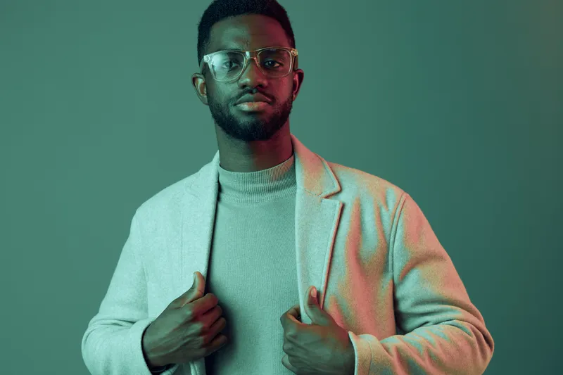 Stylish African man in a modern outfit, wearing glasses and a grey coat, posing confidently against a green backdrop, showcasing contemporary fashion trends